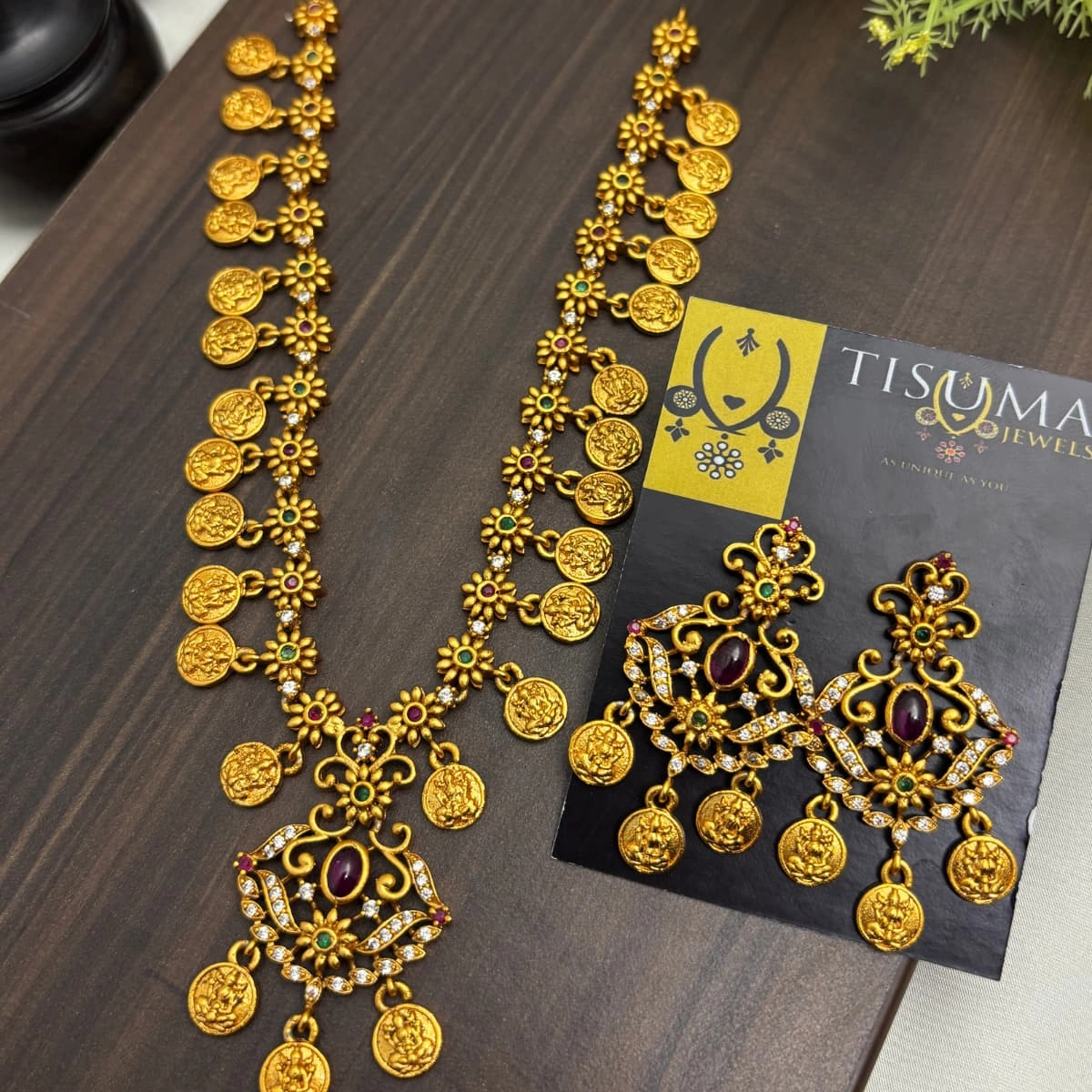 Antique gold finish Lakshmi coin necklace set with floral links, ruby and green stones, and matching coin drop earrings by Tisuma Jewels.